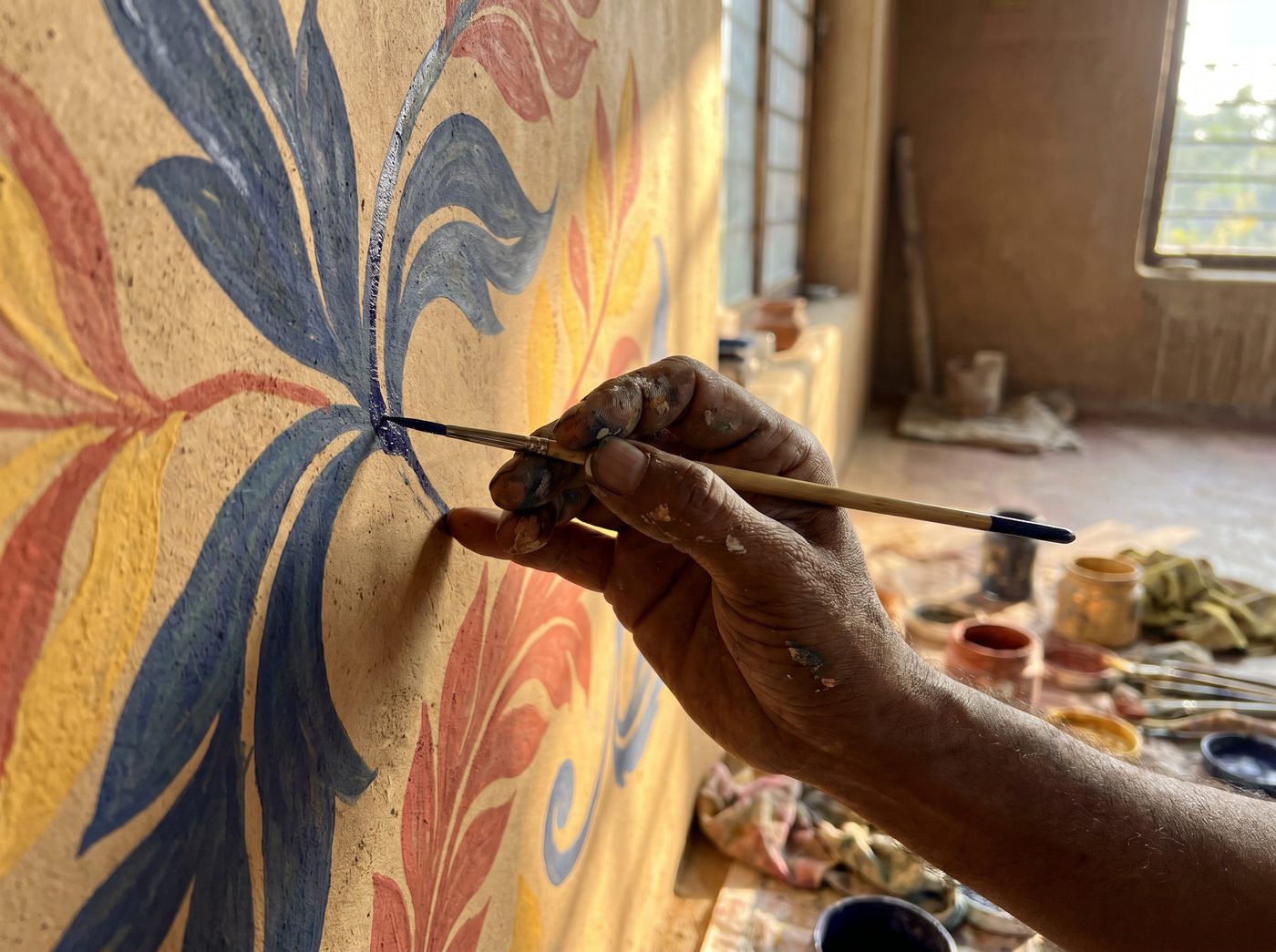 A paint-flecked hand guiding a fine brush across a half-painted indigo botanical wall